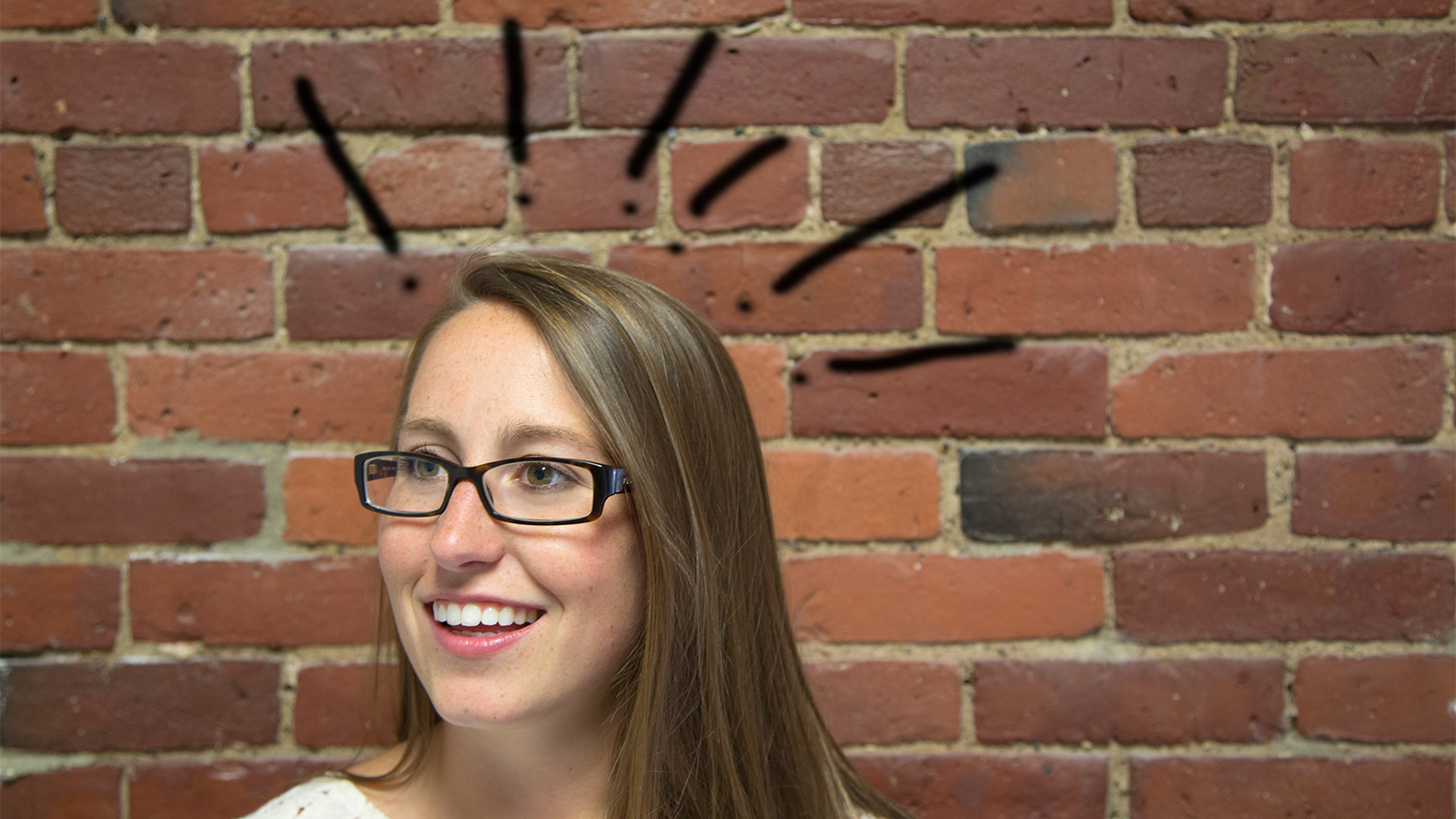 girl smiling with exclamation points above her head, she stands in front of a brick wall